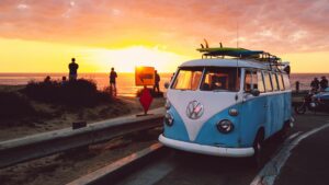 A blue and white campervan driving past the sea front and sunset with a surfboard on the roof.