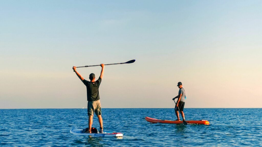 Two men paddle boarding at sea and one of them is holding their paddle in the air.