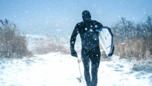 Man in a wetsuit holding a sufboard, walking through the snow.